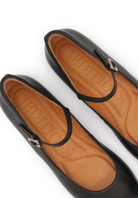 Bailarinas de cuero negro con punta redonda, que presentan una correa delgada y herrajes plateados. Forro interior color tostado con sutiles detalles de costura.