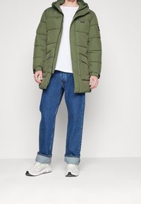 Mand iført en grøn polstret parka, hvid t-shirt, blå opsmøgede jeans og hvide sneakers, stående mod en ensfarvet baggrund.