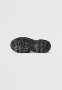 Black rubber shoe sole with rugged tread pattern, featuring deep grooves and raised accents for grip and traction. Textured surface detail.