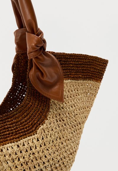 Πλεκτή τσάντα χειρός από άχυρο με σκουρόχρωμες καφέ δερμάτινες λεπτομέρειες και δερμάτινο χερούλι δεμένο με κορδόνι. Η τσάντα παρουσιάζει δίχρωμο σχέδιο με υφή.