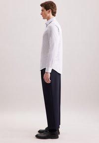 Man in profiel staand, gekleed in een lichtgestreepte blouse met lange mouwen, donkere broek en zwarte nette schoenen tegen een effen achtergrond.