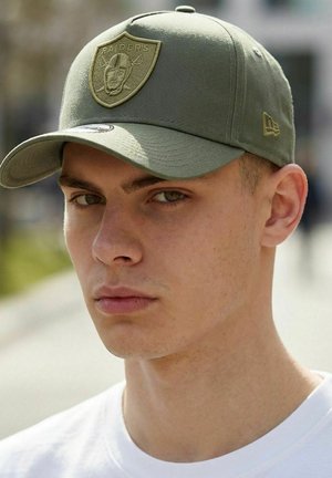 Junger Mann mit olivgrüner Raiders-Kappe mit gesticktem Logo, der nach vorne schaut, vor einem unscharfen Außenhintergrund.