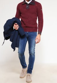 Burgunderfarbener Strickpullover mit hohem Kragen und Knopfdetails, kombiniert mit blauen Slim-Fit-Jeans und beigen Stiefeln. Eine marineblaue Jacke wird in einer Hand gehalten.
