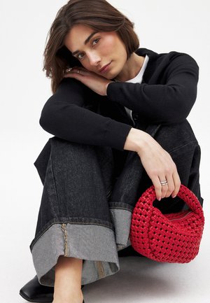 Rode geweven handtas met een ronde handgreep. Het model draagt een zwarte cardigan en donkere denim jeans met wijde pijpen, met een lichtgrijze zoom.