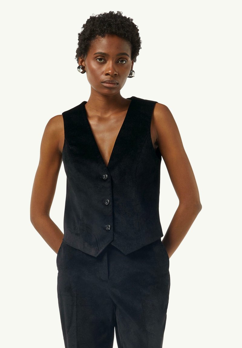 Schwarze Weste mit tiefem V-Ausschnitt, ärmellosem Design und fünf Knöpfen. Hergestellt aus weichem, strukturiertem Stoff, kombiniert mit schwarzen Hosen.