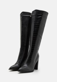Bottes noires montantes jusqu'aux genoux, en cuir synthétique texturé effet crocodile. Bout pointu avec un talon bloc robuste et une fermeture éclair latérale.