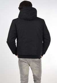 Veste à capuche noire avec une texture lisse et des poignets ajustés, assortie à un jean gris clair avec des coutures diagonales sur les poches.