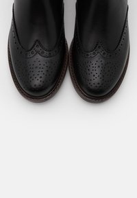 Botas de tobillo de cuero negro con un diseño brogue que presenta detalles perforados, bordes ondulados y una suela marrón. Textura suave en toda la superficie.