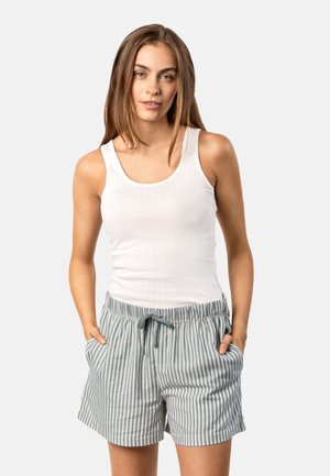 Junge Frau trägt ein weißes Tanktop und grauweiß gestreifte Shorts mit Kordelzug, steht mit den Händen in den Taschen.