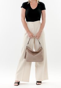 Sac à main beige avec une surface texturée et un détail à franges, porté par une personne en haut noir et pantalon large crème sur un fond uni.
