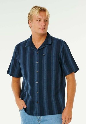 Hombre rubio con camisa azul marino de manga corta y botones, con rayas verticales, de pie con una mano en el bolsillo, mirando hacia la derecha.