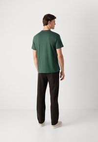 Man standing facing away, wearing a dark green short-sleeve t-shirt, black pants, and beige sneakers against a plain white background.