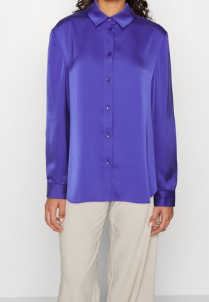 Femme portant un chemisier violet brillant à manches longues, boutonné, avec un col, associé à un pantalon beige clair, debout devant un fond uni.