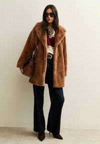 Donna con cappotto marrone in ecopelliccia oversize, maglione a righe, jeans larghi scuri, occhiali da sole e borsa marrone in mano, davanti a una parete beige.