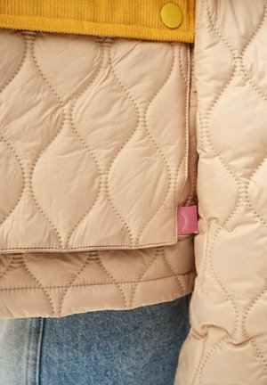 Zbliżenie na beżową pikowaną kurtkę z wzorem falistego ściegu, żółtym guzikiem oraz widoczne na dole niebieskie dżinsy.