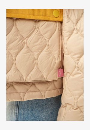 Detailní záběr na béžovou prošívanou bundu s vlnovým vzorem šití, žlutým knoflíkem a modrými džínami viditelnými dole.