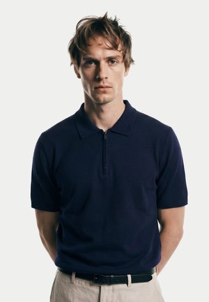 Junger Mann mit hellbraunen Haaren, der ein dunkelblaues Polohemd mit Reißverschluss und beige Hosen trägt und vor einem schlichten weißen Hintergrund steht.