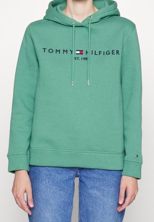 Zielona bluza z kapturem z logo "Tommy Hilfiger Est. 1985" i emblematem flagi, noszona z niebieskimi dżinsami.
