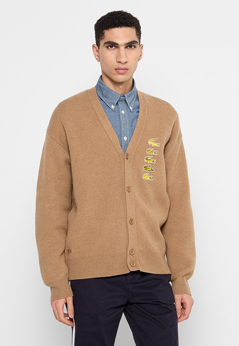 Junger Mann in einem beigen, knöpfbaren Cardigan mit gestickten gelben Krokodilen über einem blauen Hemd und schwarzen Hosen.