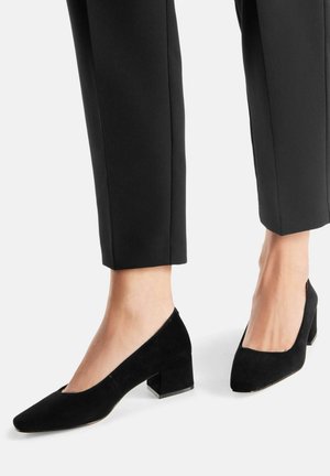 Zapatos de tacón de bloque de ante negro con punta puntiaguda y empeine bajo. Textura suave y diseño minimalista, que complementan los pantalones negros de corte ajustado.