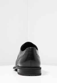 Chaussure de ville en cuir noir présentant une finition lisse, des détails brogue le long des coutures, et un talon bas avec une semelle texturée.