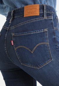 Vaqueros de mezclilla en azul oscuro con costuras amarillas; cuentan con un bolsillo trasero con una etiqueta roja y una etiqueta de cuero en la cinturilla con el logo de la marca.