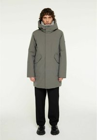 Grå vattentät parkas med huva, sidofickor och hög krage. Släthet textur och knälång design. Bärs över svarta byxor och stövlar.