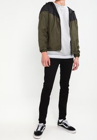 Lekka oliwkowa kurtka bomberka z czarnymi ramionami, zestawiona z szarym swetrem, czarnymi skinny jeansami i czarno-białymi sneakersami.
