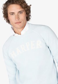 Joven con cabello rizado que lleva un suéter ligero azul con el texto "HARPER" sobre una camisa blanca, contra un fondo blanco.