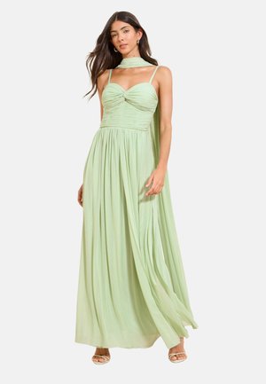 Vrouw in een vloerlang lichtgroen galajurk met spaghettibandjes en een bijpassende sjaal om haar hals, met witte hakken aan.