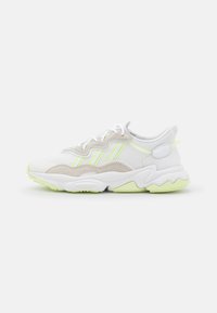 Zapatillas de malla blanca y ante, con acentos en verde neón, una suela gruesa con patrones en forma de ola y cordones.