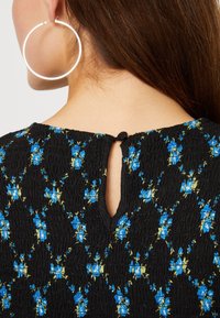 Mustat tekstiilifabric-toppi, jossa on sinisiä kukkaniittejä ja pieni avaus takana. Näkyvät suuret hopeiset rengaskorvakorut.