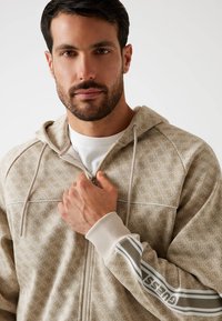 Chaqueta con capucha beige con un patrón texturizado, cierre de cremallera y puños de contraste. Presenta un logo en la manga y capucha con cordón.