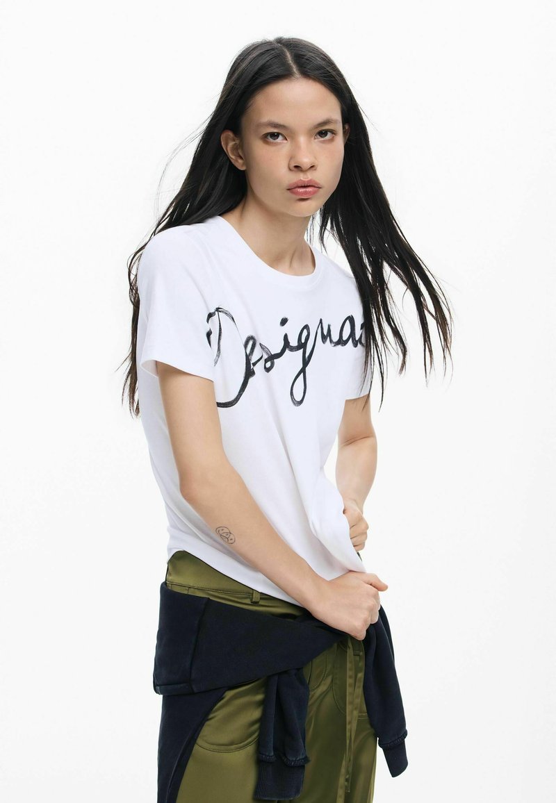 T-shirt en coton blanc avec inscription cursive noire "Design". Le modèle porte un pantalon vert olive, avec un pull bleu marine noué autour de la taille.