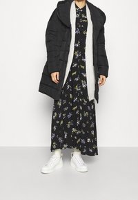 Giubbotto piumino nero con un ampio colletto, cardigan grigio a maglia sotto, e un abito maxi floreale nero. Sneakers bianche con suole testurizzate.