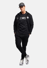 Zwarte ritssluiting hoodie met logo, zwart t-shirt met grafisch ontwerp, zwarte cargo broek en gemusterde hoge sneakers. Casual sportieve uitstraling.