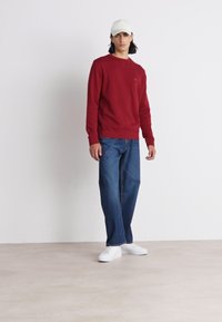 Felpa rossa con colletto a girocollo, abbinata a jeans blu larghi e sneakers bianche. Il modello indossa un cappellino bianco. Outfit semplice e casual.