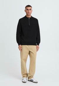 Schwarzer Zip-Kragen-Pullover mit gerippten Bündchen und Saum, kombiniert mit beigen geraden Hosen und grau-weißen Sneakers mit schwarzen Streifen.
