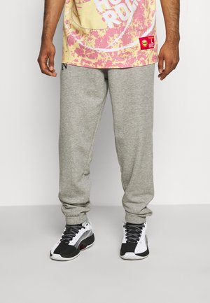 Mann trägt graue Jogginghose, schwarz-weiße Sneakers und ein gelb-pinkes Grafik-T-Shirt mit Text und rotem Label.