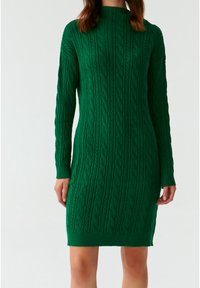 Donna che indossa un vestito in maglia a trecce verde, lungo fino al ginocchio e con maniche lunghe, su uno sfondo bianco semplice.