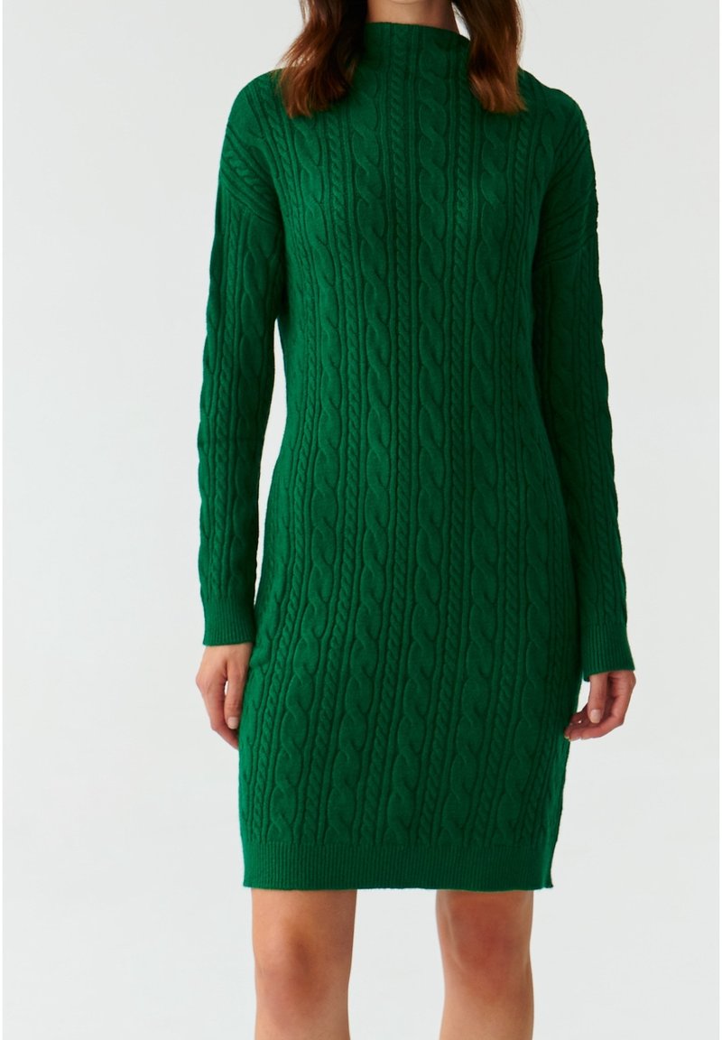 Donna che indossa un vestito in maglia a trecce verde, lungo fino al ginocchio e con maniche lunghe, su uno sfondo bianco semplice.