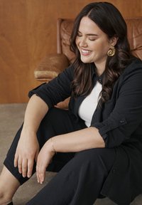 Femme aux longs cheveux foncés et boucles d'oreilles en or, assise par terre, souriant les yeux fermés, vêtue de vêtements noirs et d'un haut blanc, devant une chaise marron.