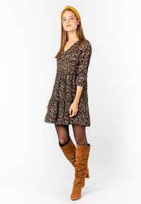 Dunkles Blumenkleid mit V-Ausschnitt und Dreiviertelärmeln, kombiniert mit braunen Wildleder-Stiefeln bis zum Knie. Es hat eine geraffte Taille und einen ausgestellten Rock.