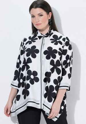 Mujer con blusa blanca con grandes estampados florales negros, mangas tres cuartos y pantalones negros sobre un fondo blanco liso.