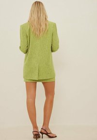 Blazer verde texturizado combinado con una falda corta a juego; cuenta con una abertura trasera sencilla y mangas largas, complementado con sandalias de tacón marrón.