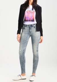 Schwarze Jacke über einem weißen T-Shirt mit grafischem Aufdruck in buntem Blumenmuster, kombiniert mit hellblauen, verwaschenen Skinny Jeans und weißen Schuhen.
