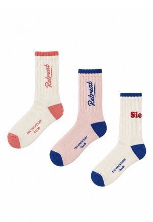 Trois chaussettes côtelées à tige mi-haute, crème et rose pâle, avec des pointes, des talons et des bords-côtes rouges et bleus contrastants, ainsi qu’un texte imprimé.