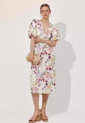 Frau in einem weißen, geblümten Midikleid mit Puffärmeln und tiefem V-Ausschnitt, hält eine beige Clutch und trägt goldene Keilsandalen.