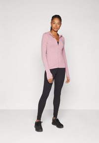 Ljusrosa figurnära zip-up jacka, lång ärm med tumhål, parat med svarta leggings och svarta träningsskor. Slät textur, minimalistisk design.