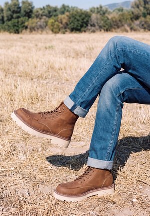 Une personne assise porte des bottes marron à lacets et un jean bleu aux revers retroussés dans un champ sec. Une des bottes est relevée.
